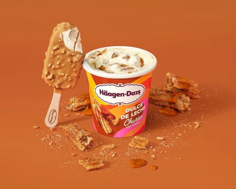 Haagen-Dazs Dulce de Leche churro ice cream pint and bar on a solid tan backdrop with crumbled churro pieces scattered around.