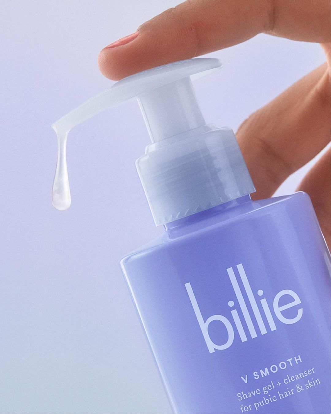 Close-up of hand dispensing Billie shave gel and cleanser from a light purple bottle with white text and pump against a light background.