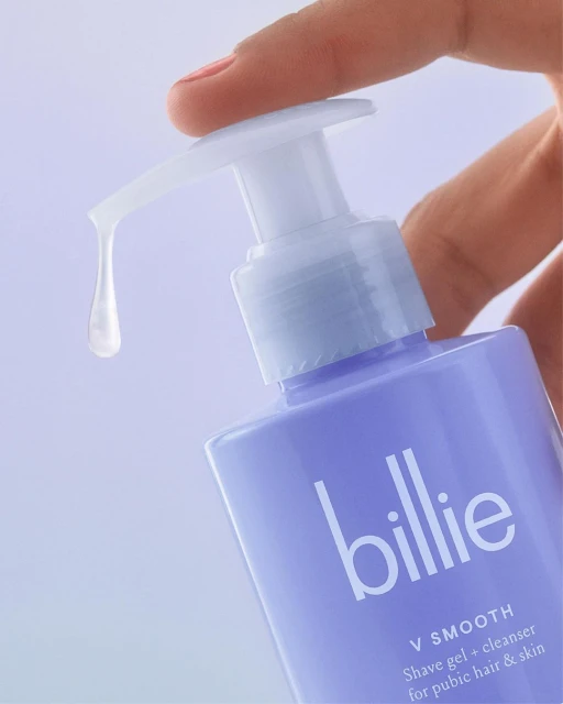 Close-up of hand dispensing Billie shave gel and cleanser from a light purple bottle with white text and pump against a light background.