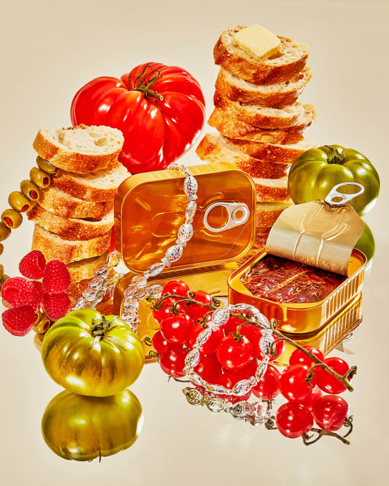 Artistic still life featuring fresh heirloom tomatoes, canned fish, sliced bread with butter, raspberries, and olives in a studio setting.