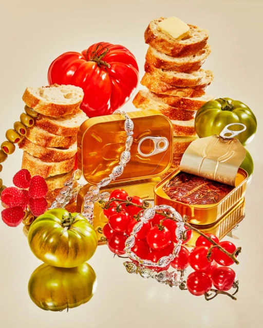 Artistic still life featuring fresh heirloom tomatoes, canned fish, sliced bread with butter, raspberries, and olives in a studio setting.