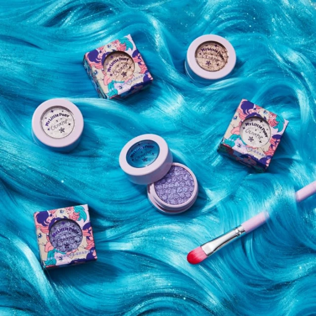 Close-up of ColourPop My Little Pony eyeshadow palettes and makeup brush on wave-patterned turquoise hair backdrop for beauty product shot.