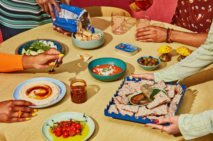 Overhead shot of various colorful food platters and bowls arranged on a yellow tablecloth, surrounded by people at a table.