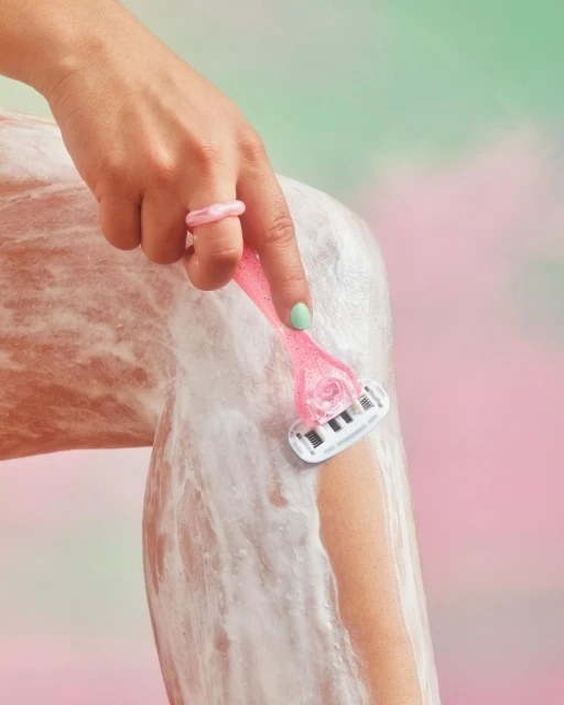 Close-up of fair-skinned woman shaving her leg with a pink razor against a pink and green pastel background, showcasing skincare.