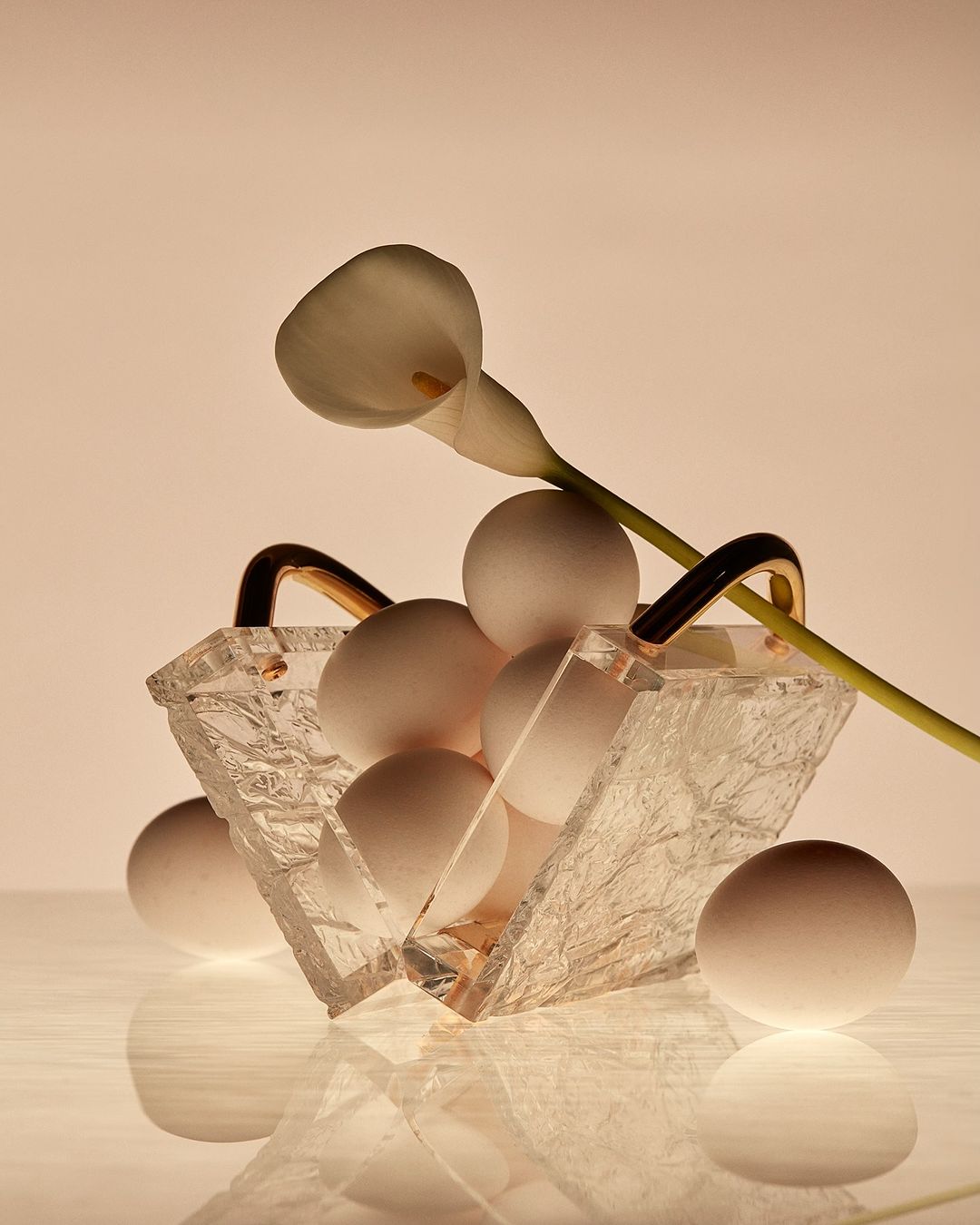 Still life composition of white eggs in a clear glass basket, adorned with a single white calla lily on a reflective surface.