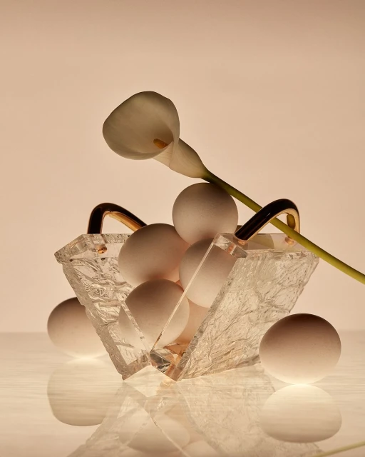 Still life composition of white eggs in a clear glass basket, adorned with a single white calla lily on a reflective surface.