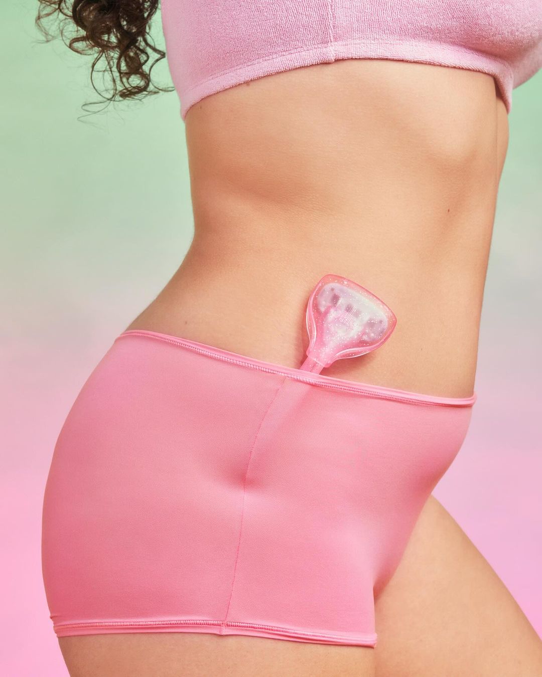 Close-up studio shot of a woman's torso and hip wearing pink shorts, holding a pink menstrual cup against a gradient background.