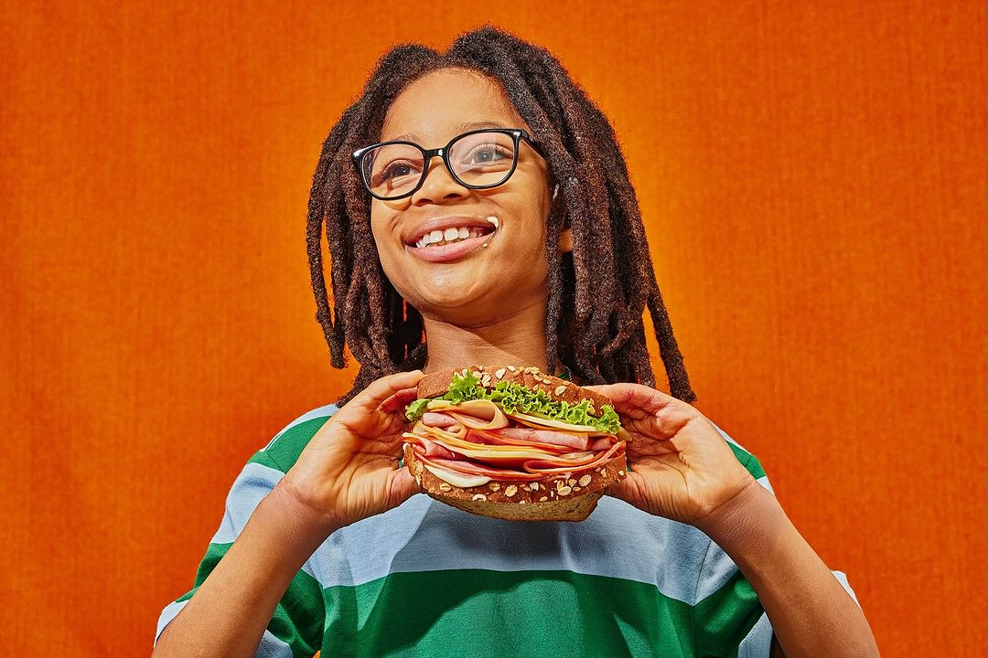 Young African-American person with dreadlocks and glasses smiles holding a ham and cheese sandwich against an orange backdrop.