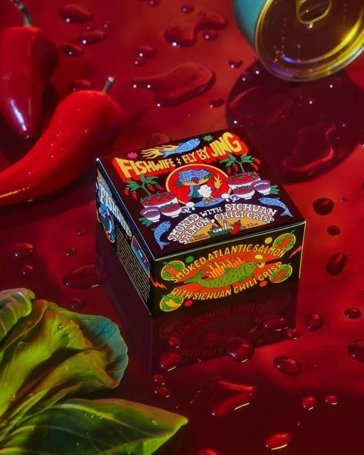 Eye-level studio shot of a Fishwife & Fly By Jing smoked salmon chili crisp box with red peppers, a green leaf, and water droplets.
