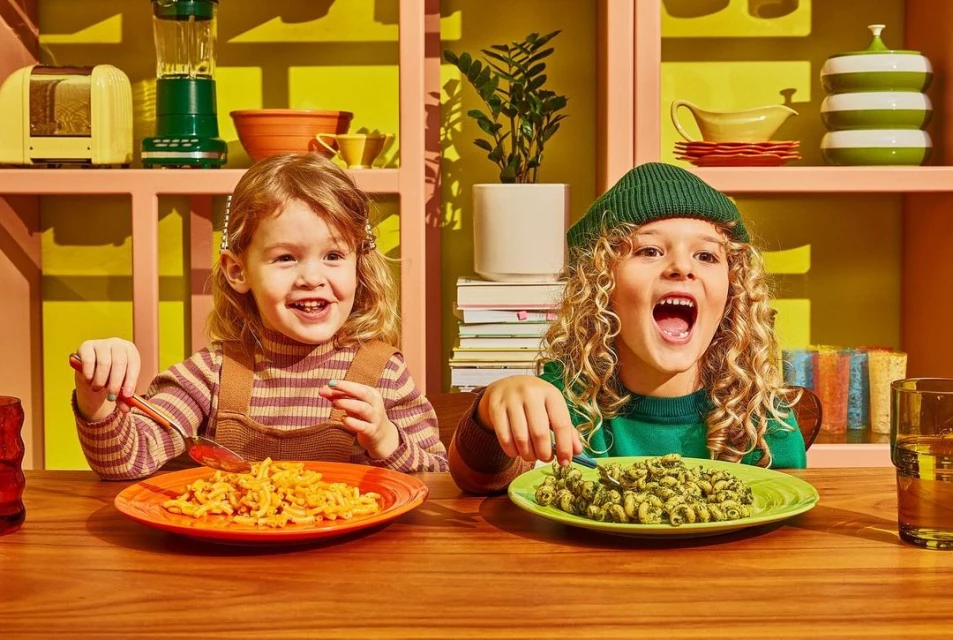 Two smiling children, a girl and a boy, are sitting at a table and eating pasta from colorful plates in a home kitchen setting.
