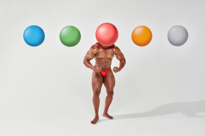 Muscular adult male with bronzed skin and red briefs posing with flexed arms, head replaced by red ball, with colored balls.