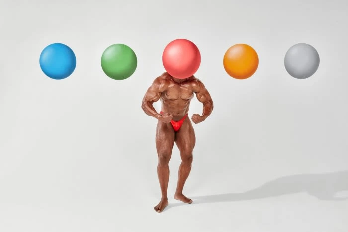 Muscular adult male with bronzed skin and red briefs posing with flexed arms, head replaced by red ball, with colored balls.