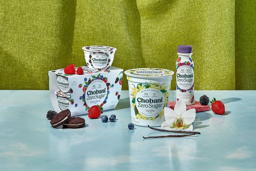Chobani Zero Sugar yogurt products in cups and bottles, assorted fresh berries, cookies, and white orchids against a green textured backdrop.