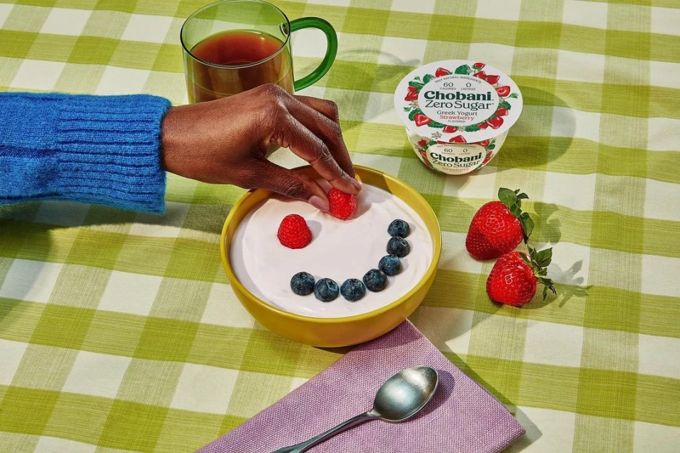 A person adds raspberries to a smiley face made of blueberries in a yellow bowl of yogurt, with Chobani yogurt and a teacup nearby.