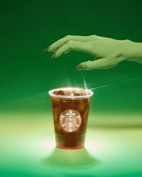 Iced coffee in a plastic cup with a green tinted hand reaching down, against a gradient green background with the Starbucks logo on the cup.
