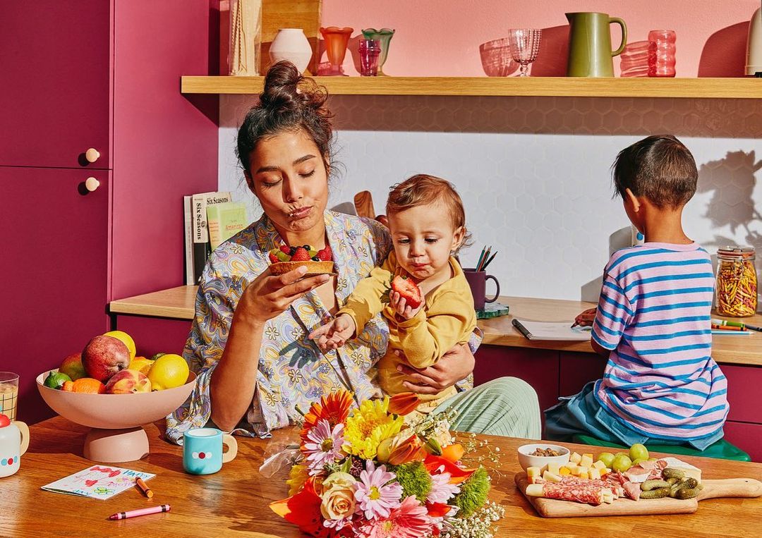 Woman and child eat tart and strawberries in a domestic kitchen with colorful decor while a boy draws at the table.