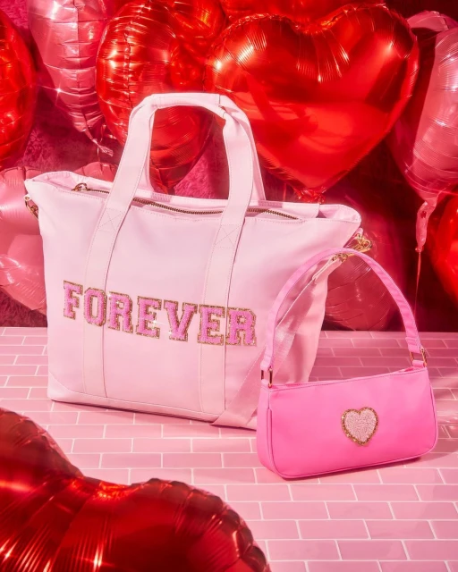 Light pink tote bag with 'Forever' text, alongside a smaller bright pink purse with a heart, against a backdrop of red heart balloons.