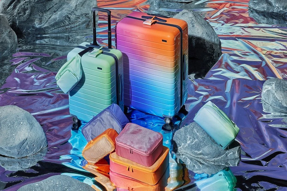 Two colorful suitcases and travel packing cubes are arranged on a shiny, reflective surface surrounded by grey rocks in a studio setting.