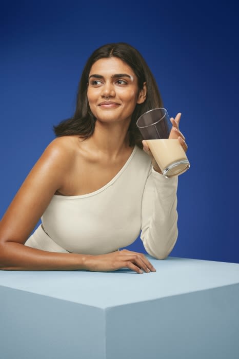 A woman in a cream-colored one-shoulder top is holding a glass with beige liquid against a blue backdrop, presented on a light blue cube.