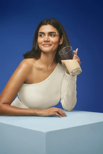 A woman in a cream-colored one-shoulder top is holding a glass with beige liquid against a blue backdrop, presented on a light blue cube.
