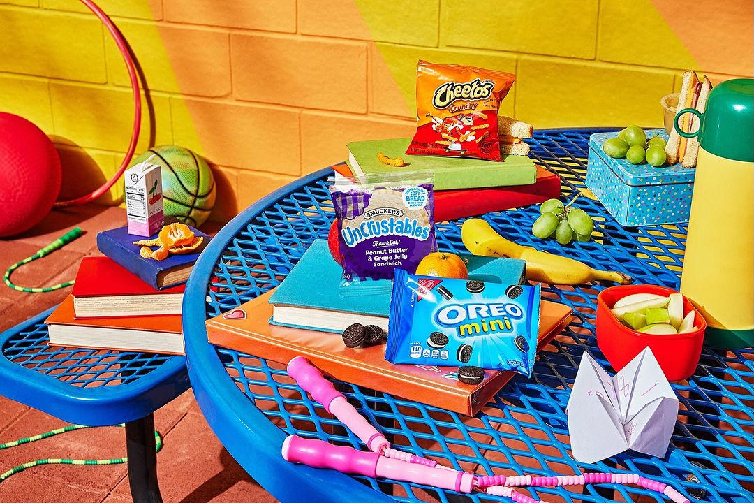 Still life of snacks and school lunch items on a blue picnic table with a yellow and orange background, arranged on colorful books.