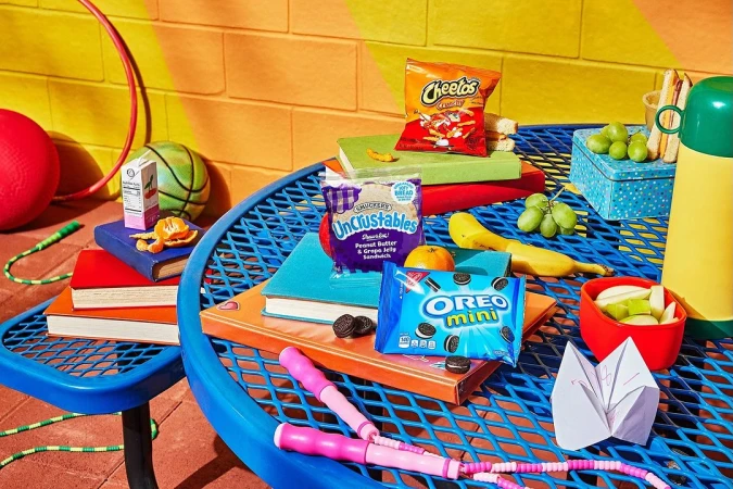 Still life of snacks and school lunch items on a blue picnic table with a yellow and orange background, arranged on colorful books.