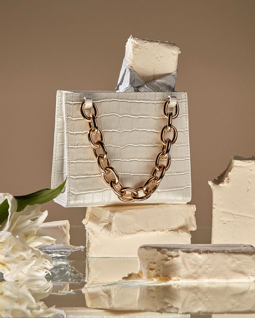 White faux crocodile purse with gold chain strap displayed on a stack of frosted white blocks, next to a white floral arrangement. 