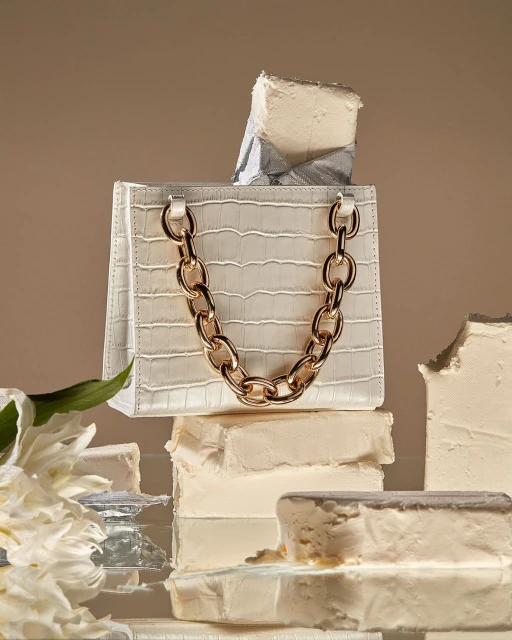White faux crocodile purse with gold chain strap displayed on a stack of frosted white blocks, next to a white floral arrangement. 