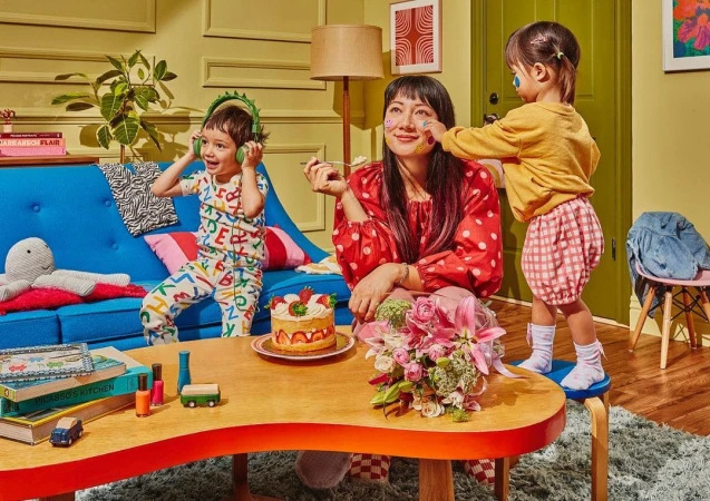 Woman and two children in playful scene with cake and paint, set indoors with blue sofa, plants, and colorful decor.