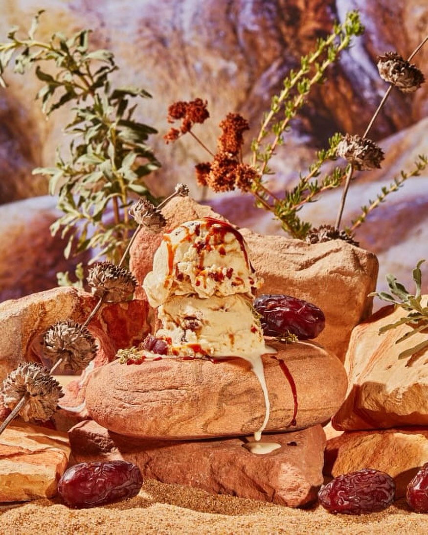 Ice cream scoops with caramel drizzle are arranged on rock formations, surrounded by natural desert vegetation and dates.