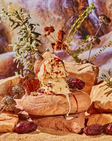Ice cream scoops with caramel drizzle are arranged on rock formations, surrounded by natural desert vegetation and dates.
