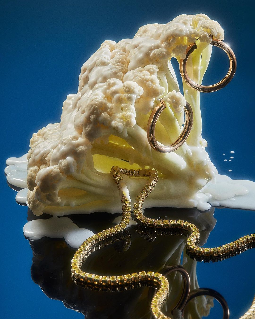 Still life photograph featuring a head of cauliflower adorned with gold hoop earrings and a gold chain necklace against a gradient blue background.