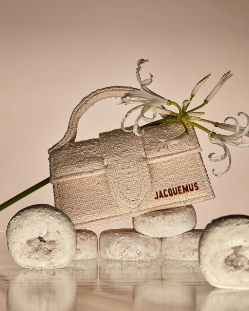 Jacquemus handbag on stack of powdered donuts with white amaryllis flowers in a studio setting with warm lighting. Fashion product shot.