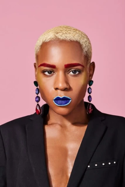 Portrait of a short blonde haired person in a black jacket with blue lipstick, red eyebrows, and gemstone earrings against pink.