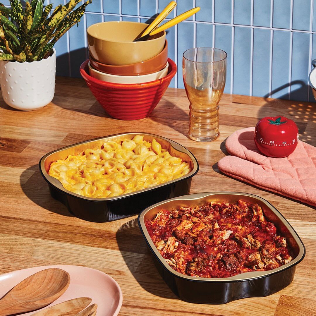 Two trays of cooked food, mac and cheese and another with red sauce, sit on a wooden counter next to dishes, a glass, and a timer.