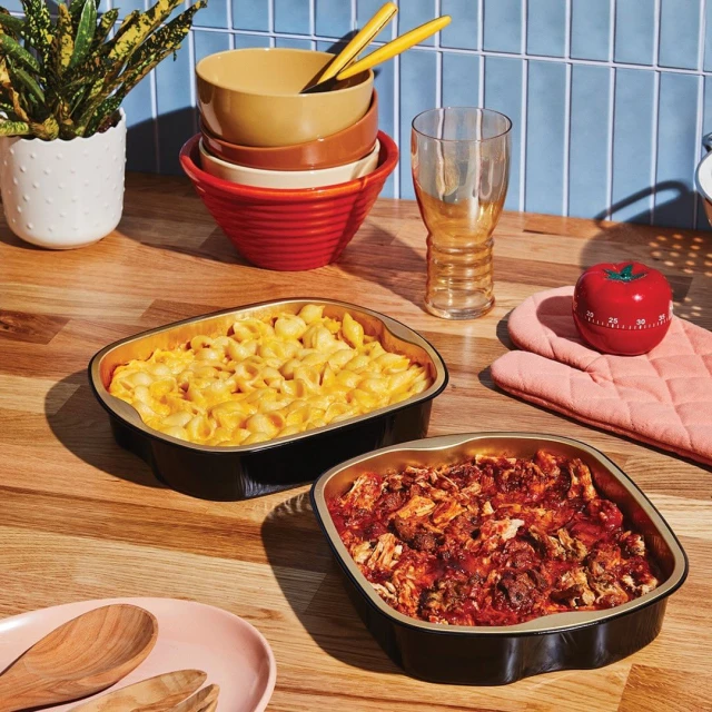 Two trays of cooked food, mac and cheese and another with red sauce, sit on a wooden counter next to dishes, a glass, and a timer.