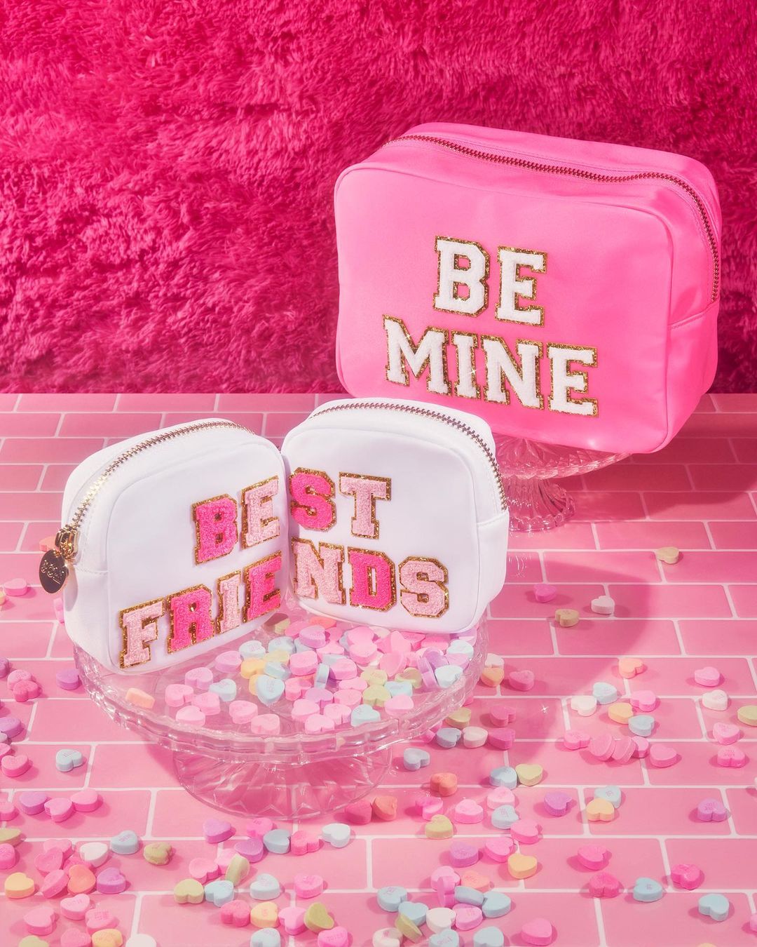 Three cosmetic bags are arranged on a pink brick surface with candy hearts scattered around them against a bright pink background.