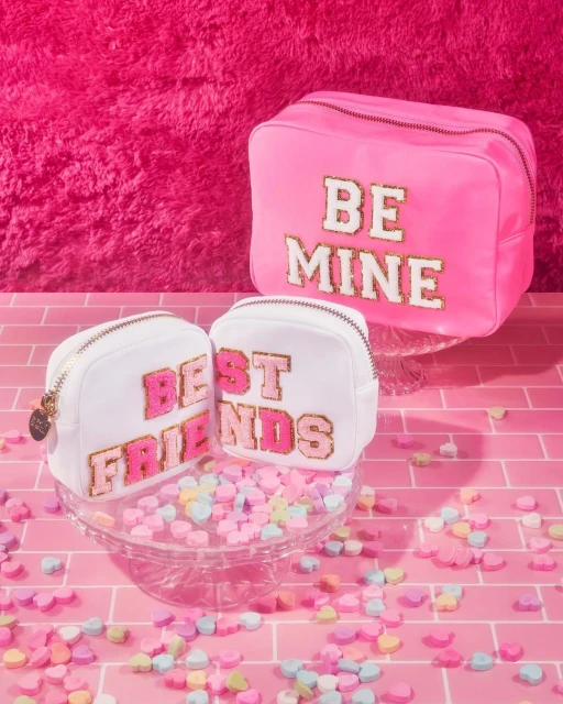 Three cosmetic bags are arranged on a pink brick surface with candy hearts scattered around them against a bright pink background.