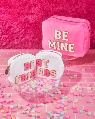 Three cosmetic bags are arranged on a pink brick surface with candy hearts scattered around them against a bright pink background.