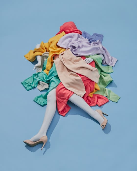 Fashion still life on a blue background featuring a mannequin torso and legs draped in colorful sweaters and skirts with beige heels.