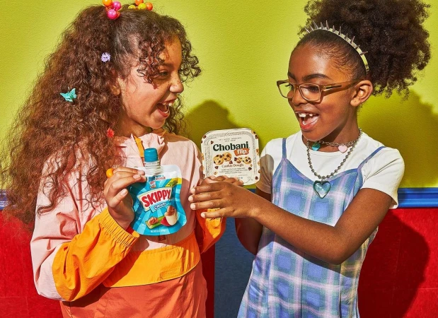 Two smiling young girls indoors holding a Chobani Flip yogurt and a Skippy peanut butter pouch against a brightly coloured background.