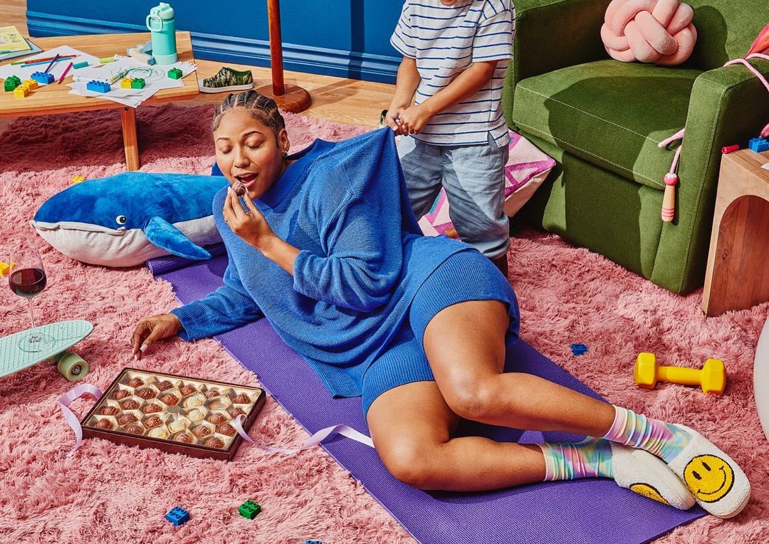 Woman in blue loungewear eats a chocolate while lying on a yoga mat, with a child grabbing her top and toys around her in a room.