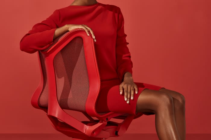 A person in a red dress sits in a red office chair against a matching red background, creating a monochromatic and visually striking image.