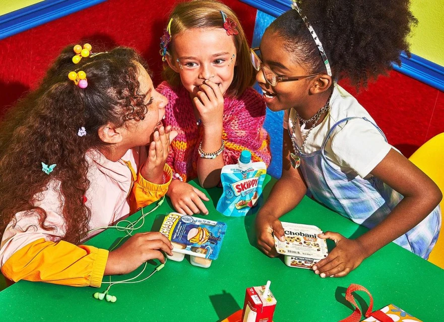 Three young girls share a snack at a green table against a vibrant, colorful background, enjoying Chobani yogurt and Skippy peanut butter.