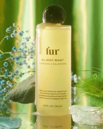 Bottle of Fur All Body Wash on a reflective surface with green plants and blue flowers in the background. Beauty product shot.