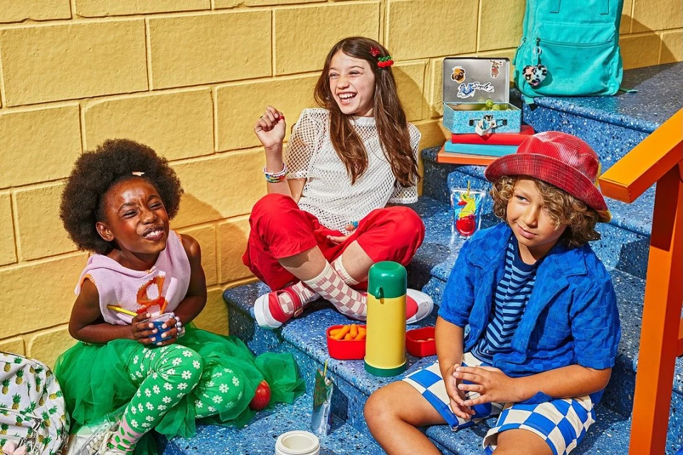 Three happy children sit on blue steps, sharing snacks and drinks from lunchboxes, creating a candid, lifestyle image with a yellow brick wall background.