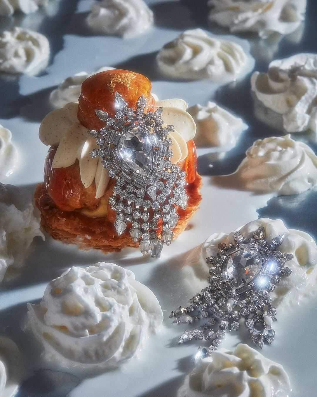 Cream puff pastry with decorative jewel brooches, surrounded by dollops of whipped cream on a light blue-grey surface in studio light.