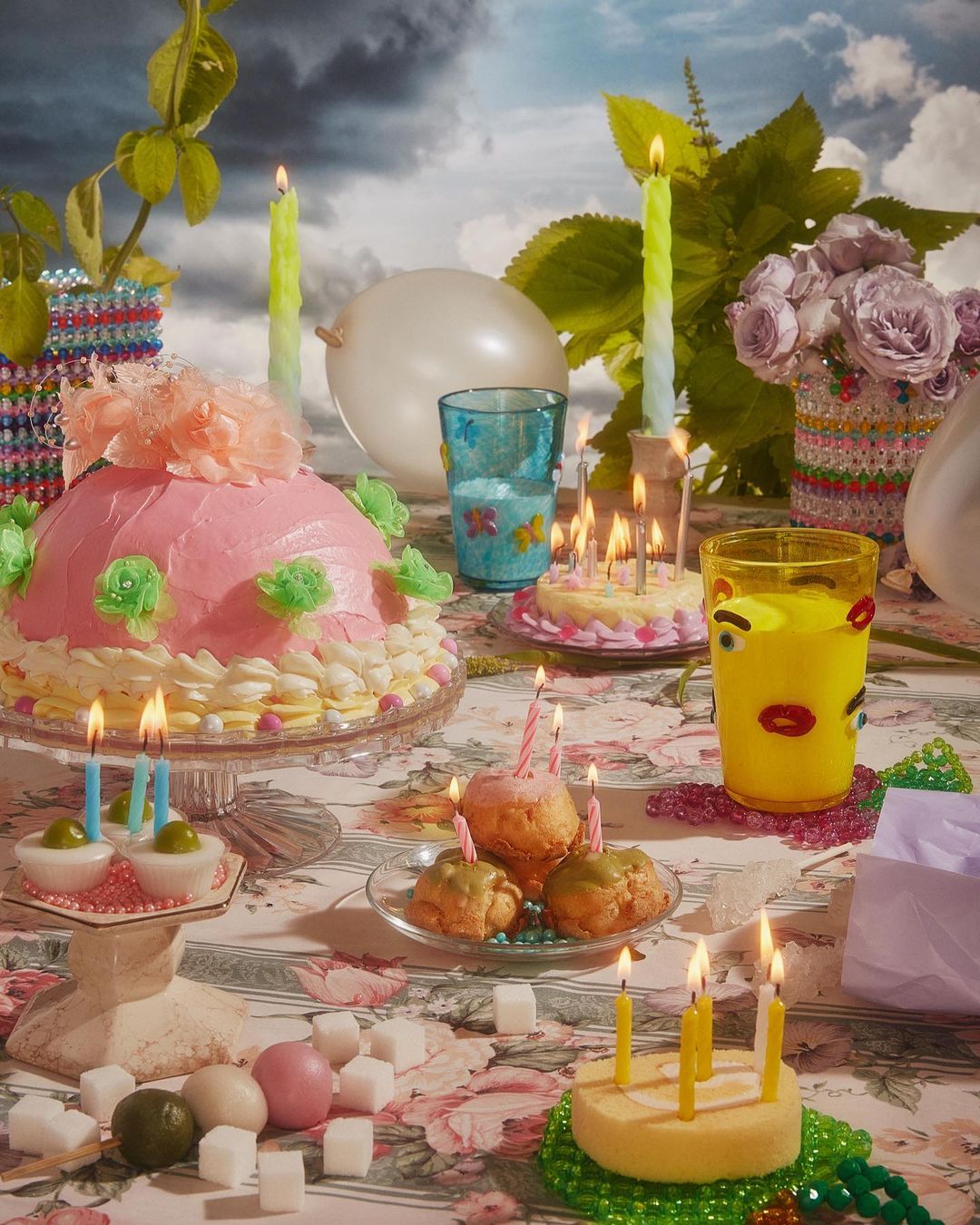 A table with a pink cake, lit candles, glass cups, flowers, sugar cubes and eggs against a cloudy sky backdrop. Party theme.