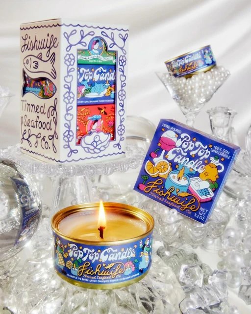 Collection of Pop Top Candle tins and packaging by Fishwife Tinned Seafood Co, featuring vibrant designs, stacked on reflective surface.