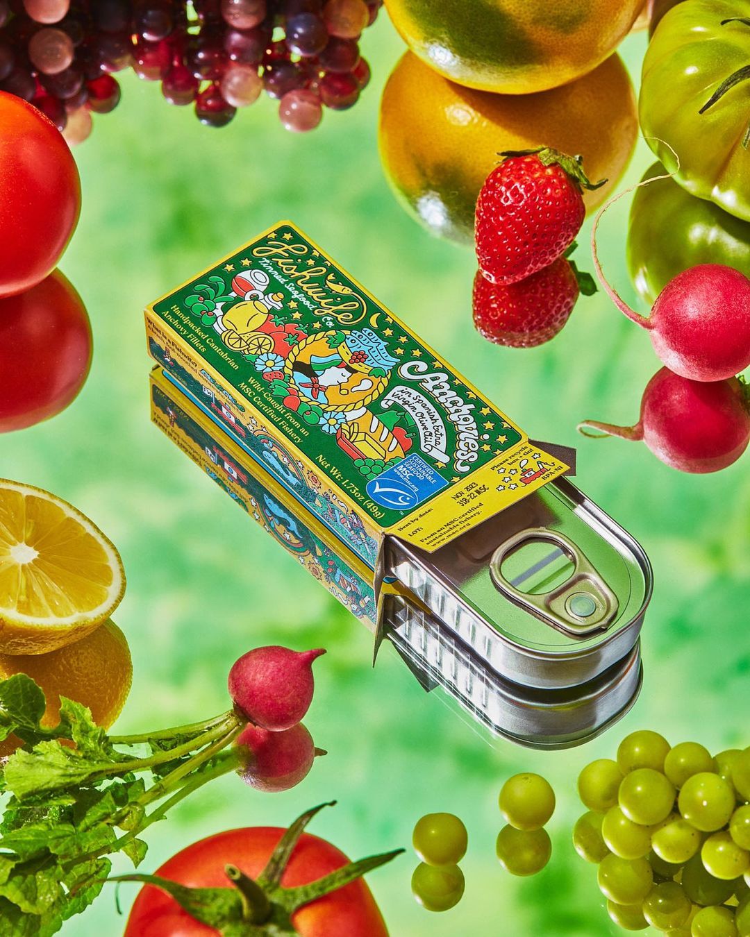 Artistic food still life featuring an open tin of anchovies, tomatoes, lemons, grapes, and radishes on a glossy green surface.